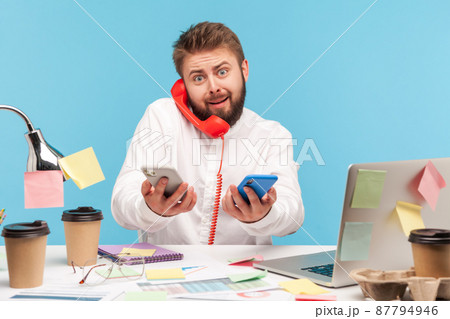 Scared man office worker answering call at workplace all covered with sticky notes and holding mobile phones in hands, service centre, technical support. Indoor studio shot isolated on blue background Scared man office worker answering call at workplace all covered with sticky notes and holding mobile phones in hands, service centre, technical support. Indoor studio shot isolated on blue background 87794946