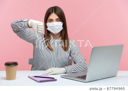 Displeased woman employee wearing hygienic mask and protective gloves during quarantine, showing thumbs down, dislike gesture. Working at home office in self-isolation, coronavirus outbreak. Indoor 87794960