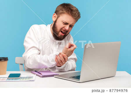 Bearded man office worker in white shirt grimacing feeling tingling, numbness and pain in wrist working on laptop, suffering carpal tunnel syndrome. Indoor studio shot isolated on blue background 87794976