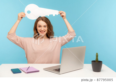 Positive emotions about property purchase, rental office. Amazing woman sitting at workplace with laptop and holding big paper key over head, pleased with real estate agency service. indoor isolated 87795005