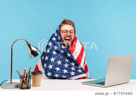 Extremely happy funny man employee sitting wrapped in American flag and shouting for joy in office workplace, celebrating labor day or US Independence day 4th of july, government employment support. 87795080