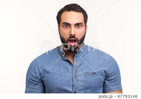 Surprised wondered man with beard in blue shirt looking at camera with big eyes and open mouth, shocked with unexpected news. Indoor studio shot isolated on white background Surprised wondered man with beard in blue shirt looking at camera with big eyes and open mouth, shocked with unexpected news. Indoor studio shot isolated on white background 87795146