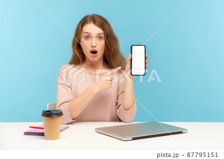 Surprised astonished woman, office employee, sitting at workplace and pointing cellphone mock up display, copy space for application, website advertise. indoor studio shot isolated on blue background 87795151