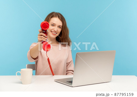Positive call center operator, woman secretary sitting at workplace and holding old fashioned phone handset, smiling friendly, asking to answer phone. indoor studio shot isolated on blue background Positive call center operator, woman secretary sitting at workplace and holding old fashioned phone handset, smiling friendly, asking to answer phone. indoor studio shot isolated on blue background 87795159