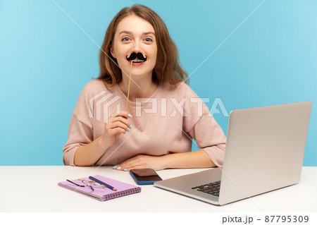 Funny woman office employee covering lips with fake paper moustache, imitating male boss and smiling joyfully, wearing humorous masquerade accessory. indoor studio shot isolated on blue background 87795309