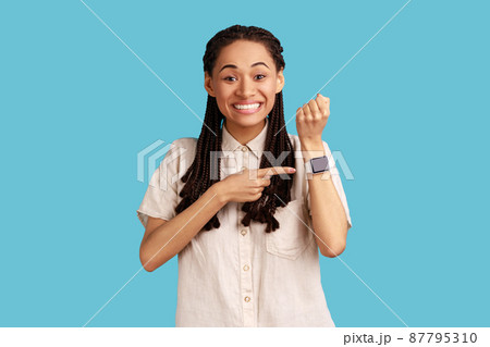 Portrait of happy excited woman with black dreadlocks standing, looking at camera, pointing at her smart watch, wearing white shirt. Indoor studio shot isolated on blue background. 87795310