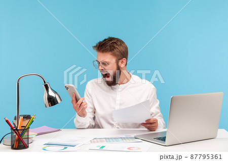 Angry nervous man office worker in white shirt screaming looking at smartphone display, skipping important event through lot of paper work. Indoor studio shot isolated on blue background Angry nervous man office worker in white shirt screaming looking at smartphone display, skipping important event through lot of paper work. Indoor studio shot isolated on blue background 87795361
