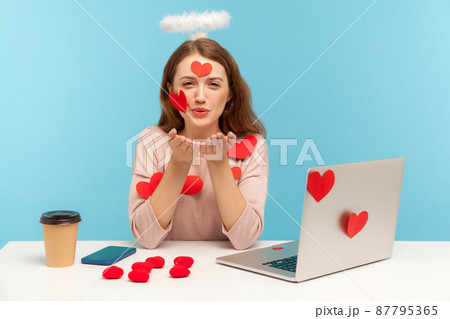 Angelic woman with kind expression sitting at workplace office, all covered with sticker love hearts, sending romantic sensual air kiss, full of valentine's day greetings. indoor studio shot, isolated 87795365