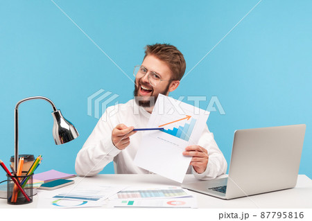 Happy bearded man in eyeglasses smiling at camera pointing at growth diagram, satisfied with financial and economic growth of his business. Indoor studio shot isolated on blue background Happy bearded man in eyeglasses smiling at camera pointing at growth diagram, satisfied with financial and economic growth of his business. Indoor studio shot isolated on blue background 87795816