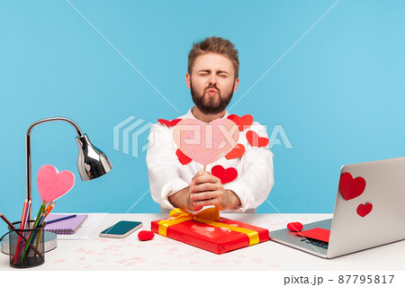 Funny bearded man sitting at workplace all covered with sticky hearts and sending air kisses holding pink heart greeting card, making gift. Indoor studio shot isolated on blue background Funny bearded man sitting at workplace all covered with sticky hearts and sending air kisses holding pink heart greeting card, making gift. Indoor studio shot isolated on blue background 87795817