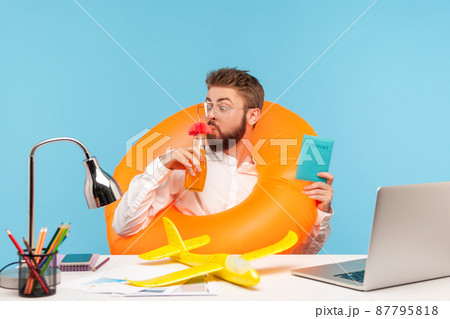 Funny happy man office worker drinking lemonade sitting in rubber lifebuoy and holding passport, going to fly in warm countries, planning vacation. Indoor studio shot isolated on blue background Funny happy man office worker drinking lemonade sitting in rubber lifebuoy and holding passport, going to fly in warm countries, planning vacation. Indoor studio shot isolated on blue background 87795818