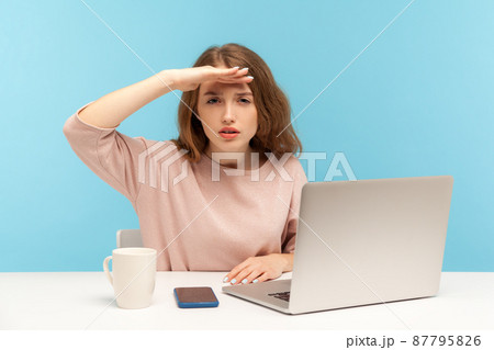 Ambitious young woman employee sitting at workplace with laptop and looking far away with hand above eyes, searching with view, exploring distant future. indoor studio shot isolated on blue background 87795826