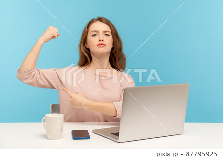 Independent strong confident woman boss sitting at workplace with laptop and showing biceps, looking proud, feeling power to success in business. indoor studio shot isolated on blue background 87795925