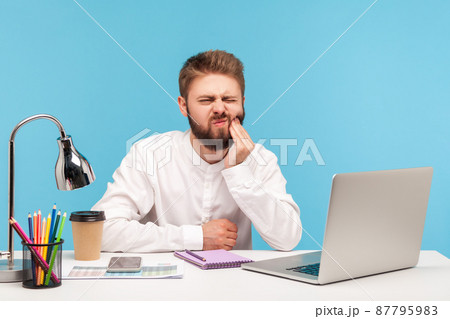 Unhappy bearded man office worker touching cheek with fingers feeling unpleasant toothache, suffering gums inflammation, dentistry problems. Indoor studio shot isolated on blue background 87795983