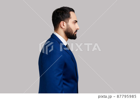 Side view portrait of confident bearded male looking at camera with serious expression, unsmiling determined business man, wearing official style suit. Indoor studio shot isolated on gray background. 87795985