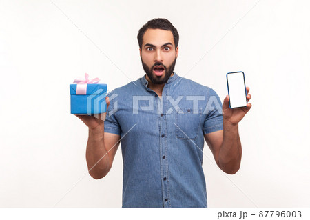 Excited surprised man with beard holding gift box and smartphone with empty screen looking at camera with big eyes, shocked with present. Indoor studio shot isolated on white background 87796003