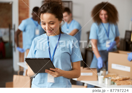 Portrait of charming young female volunteer in blue uniform using tablet pc and smiling while standing indoors. Team sorting, packing items in the background 87797224