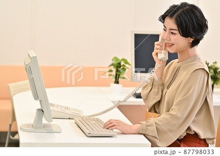 オフィスでディスクのパソコンの前で電話をする女性 87798033