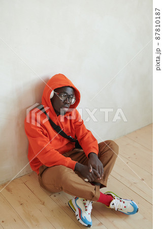 High angle studio shot of young African American man wearing stylish orange hoodie sitting on wooden floor listening to music in headphones High angle studio shot of young African American man wearing stylish orange hoodie sitting on wooden floor listening to music in headphones 87810187