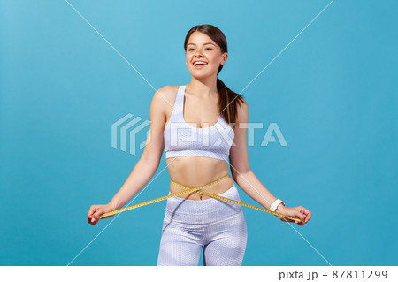 Slim athletic woman in white sports tights and top crossing tape line on her waist, measuring centimeters of body, keeping healthy lifestyle, workout. Indoor studio shot isolated on blue background Slim athletic woman in white sports tights and top crossing tape line on her waist, measuring centimeters of body, keeping healthy lifestyle, workout. Indoor studio shot isolated on blue background 87811299
