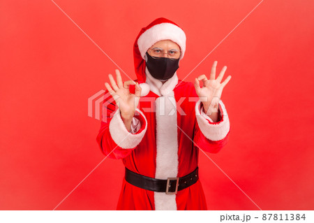 Positive optimistic man in santa claus costume with protective face mask showing ok gesture with fingers, cheering up on quarantine. Indoor studio shot isolated on red background 87811384