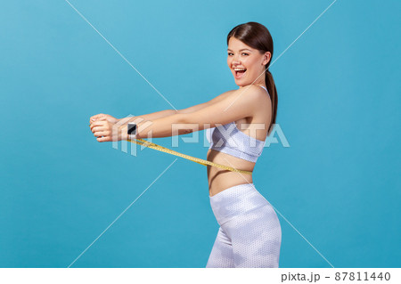 Profile portrait happy slim woman in white sportswear holding on waist tape measure and looking at camera with satisfaction and smile. Indoor studio shot isolated on blue background 87811440
