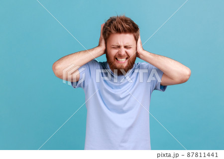 Don't want to listen. Portrait of despaired man irritated by loud noise covering ears and grimacing in pain, suffering annoying high-decibel sound. Indoor studio shot isolated on blue background. 87811441
