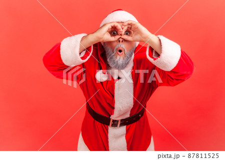 Portrait of curious funny gray bearded man in santa claus costume holding hands near eyes looking like through binocular and prying about holidays wishes. Indoor studio shot isolated on red background 87811525