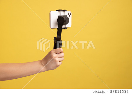 Closeup side view of woman hand holding steadicam with phone, for making video or has livestream. Indoor studio shot isolated on yellow background. Closeup side view of woman hand holding steadicam with phone, for making video or has livestream. Indoor studio shot isolated on yellow background. 87811552
