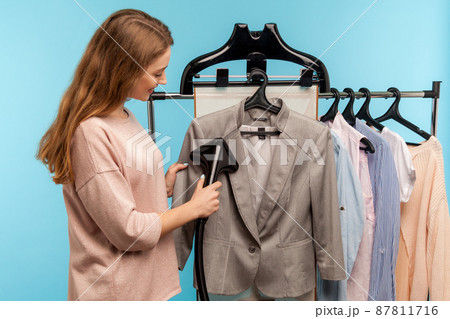 Cheerful housewife steaming business jacket at dry-cleaner and looking at fabric, advertisement of professional services washing, cleaning and ironing garment. indoor studio shot, blue background 87811716