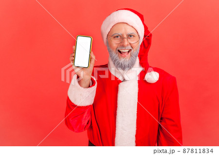 Happy gray bearded santa claus holding and showing smartphone with white display looking at camera with toothy smile. Indoor studio shot isolated on red background Happy gray bearded santa claus holding and showing smartphone with white display looking at camera with toothy smile. Indoor studio shot isolated on red background 87811834