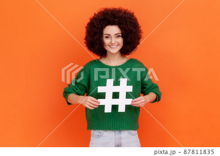 Shocked woman with Afro hairstyle pointing at white hashtag symbol, astonishing promoting viral topic in social network, tagging blog trends. Indoor studio shot isolated on orange background. Shocked woman with Afro hairstyle pointing at white hashtag symbol, astonishing promoting viral topic in social network, tagging blog trends. Indoor studio shot isolated on orange background. 87811835