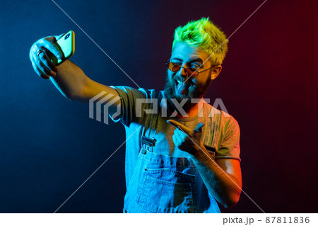 Handsome hipster bearded man wearing t shirt and denim overalls, broadcasting live stream, points to camera with finger, grimacing, winking and showing tongue. Colorful neon light, indoor studio shot Handsome hipster bearded man wearing t shirt and denim overalls, broadcasting live stream, points to camera with finger, grimacing, winking and showing tongue. Colorful neon light, indoor studio shot 87811836