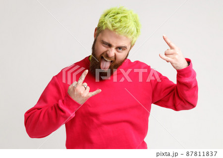 Portrait of carefree excited person showing tongue and heavy metal symbol with two arms fingers, looks at camera with cocky expression. Indoor studio shot isolated on gray background. Portrait of carefree excited person showing tongue and heavy metal symbol with two arms fingers, looks at camera with cocky expression. Indoor studio shot isolated on gray background. 87811837