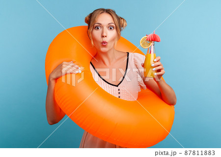 Blonde adorable woman with astonished facial expression looking directly at camera and combines lips a tubule, holding rubber ring and cocktail. Indoor studio shot isolated on blue background. Blonde adorable woman with astonished facial expression looking directly at camera and combines lips a tubule, holding rubber ring and cocktail. Indoor studio shot isolated on blue background. 87811883
