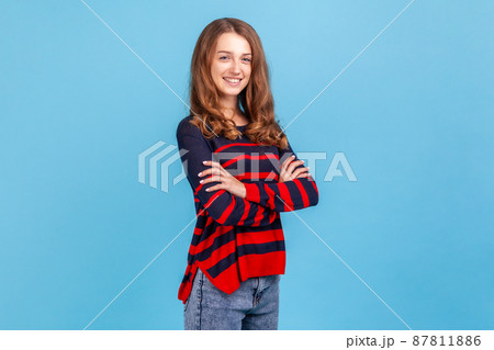 Side view of confident woman wearing striped casual style sweater standing with folded arms, looking at camera with happy expression, good mood. Indoor studio shot isolated on blue background. Side view of confident woman wearing striped casual style sweater standing with folded arms, looking at camera with happy expression, good mood. Indoor studio shot isolated on blue background. 87811886