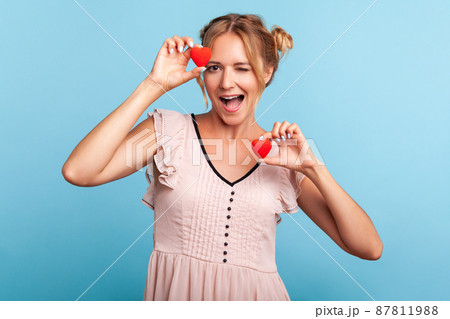 Portrait of friendly positive blonde woman in dress with two hair buns holding heart symbols in hands and winking at camera, sending love. Indoor studio shot isolated on blue background. Portrait of friendly positive blonde woman in dress with two hair buns holding heart symbols in hands and winking at camera, sending love. Indoor studio shot isolated on blue background. 87811988