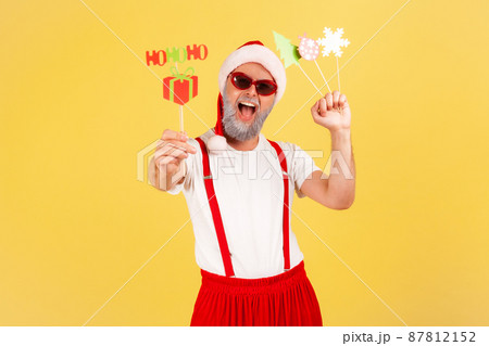 Extremely positive excited grey bearded adult man in red hat holding congratulations cards on sticks, new year and christmas celebration. Indoor studio shot isolated on yellow background 87812152