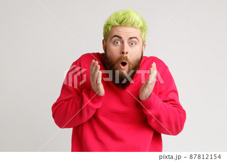 Photo of amazed shocked bearded young man with yellow hair, looks at camera with open mouth, raising palms, being impressed of shocking news. Indoor studio shot isolated on gray background. 87812154