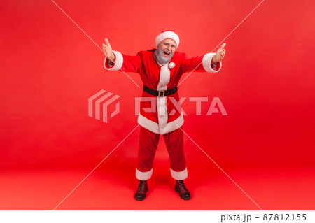 Full length portrait of smiling friendly man in santa claus costume raising hands wanting to embrace and congratulate, free hugs, magic of holidays. Indoor studio shot isolated on red background 87812155