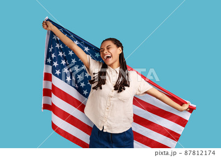 Cheerful attractive woman with black dreadlocks holding USA flag over shoulders and keeps eyes closed and smiling happily, wearing white shirt. Indoor studio shot isolated on blue background. 87812174