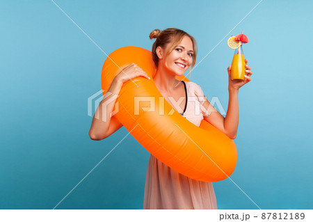Happy blonde woman with orange rubber ring rising hand with cocktail and looking directly at camera with charming smile, wearing summer dress. Indoor studio shot isolated on blue background. Happy blonde woman with orange rubber ring rising hand with cocktail and looking directly at camera with charming smile, wearing summer dress. Indoor studio shot isolated on blue background. 87812189