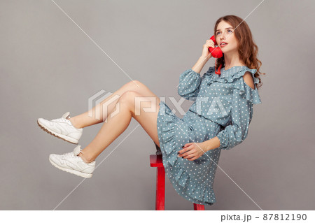 Charming girl in ruffle dress sitting delicate graceful airy on chair, holding phone handset enjoying pleasant conversation on telephone, looking up dreamy relaxed. indoor studio shot isolated on gray 87812190