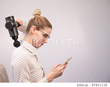 A young woman sits on a chair, uses a smartphone and makes herself massage with an electro massage gun. 87821116
