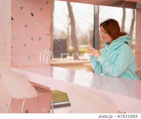 Caucasian red-haired woman eats ice cream with a plastic spoon while sitting alone in a cafe. 87831969