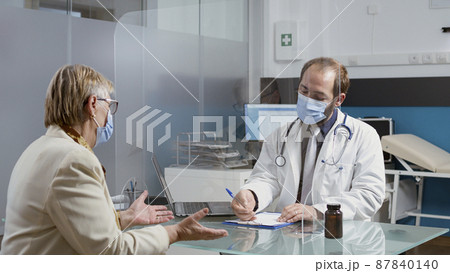 Specialist taking notes at consultation with senior patient, writing prescription treatment papers during pandemic. Medic and elder woman talking about healthcare service in cabinet. 87840140