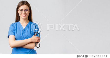Healthcare workers, medicine, insurance and covid-19 pandemic concept. Confident and enthusiastic young female nurse, doctor in blue scrubs and glasses, hold stethoscope, smiling camera pleased Healthcare workers, medicine, insurance and covid-19 pandemic concept. Confident and enthusiastic young female nurse, doctor in blue scrubs and glasses, hold stethoscope, smiling camera pleased 87845731