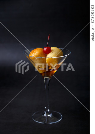 Neapolitan Rum baba (or baba au rum) in a martini glass with a cocktail cherry on a black background. Small yeast cakes soaked in rum syrup. Traditional Italian pastry. Close-up, selective focus. Neapolitan Rum baba (or baba au rum) in a martini glass with a cocktail cherry on a black background. Small yeast cakes soaked in rum syrup. Traditional Italian pastry. Close-up, selective focus. 87850533