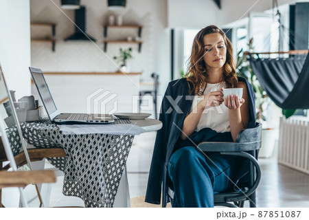 A woman works at home, sitting at a table with a laptop and holding a mug 87851087