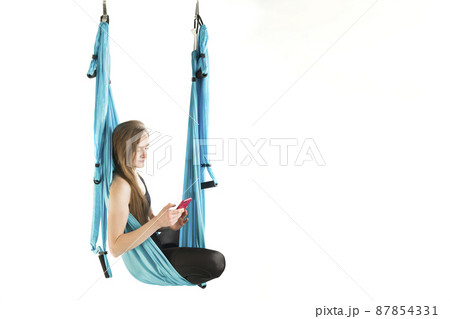 Yoga instructor has break and holding phone. Young athletic woman hang in hammock during an aerial gymnastics or fly yoga workout. Yoga instructor has break and holding phone. Young athletic woman hang in hammock during an aerial gymnastics or fly yoga workout. 87854331